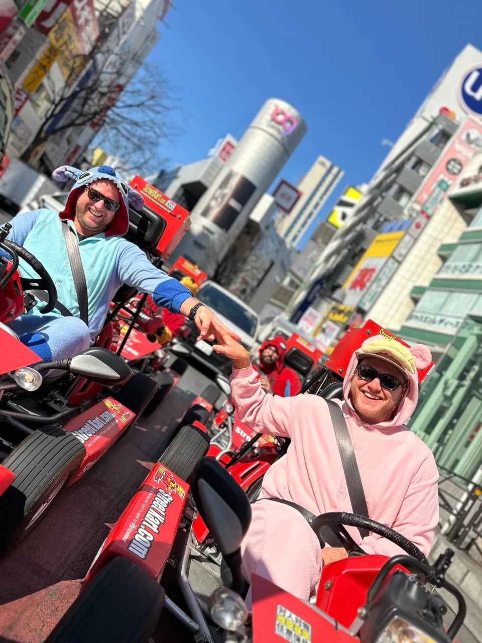 Two men in colorful outfits ride red go-karts on a busy city street, high-fiving as they race, with tall buildings and bright signs behind them.