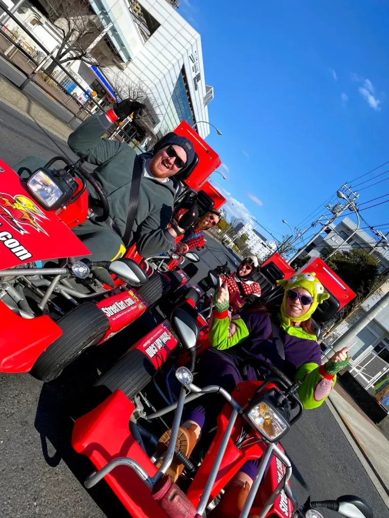 Group of friends riding bright red pedal go-karts on a sunny urban street, giving thumbs up.