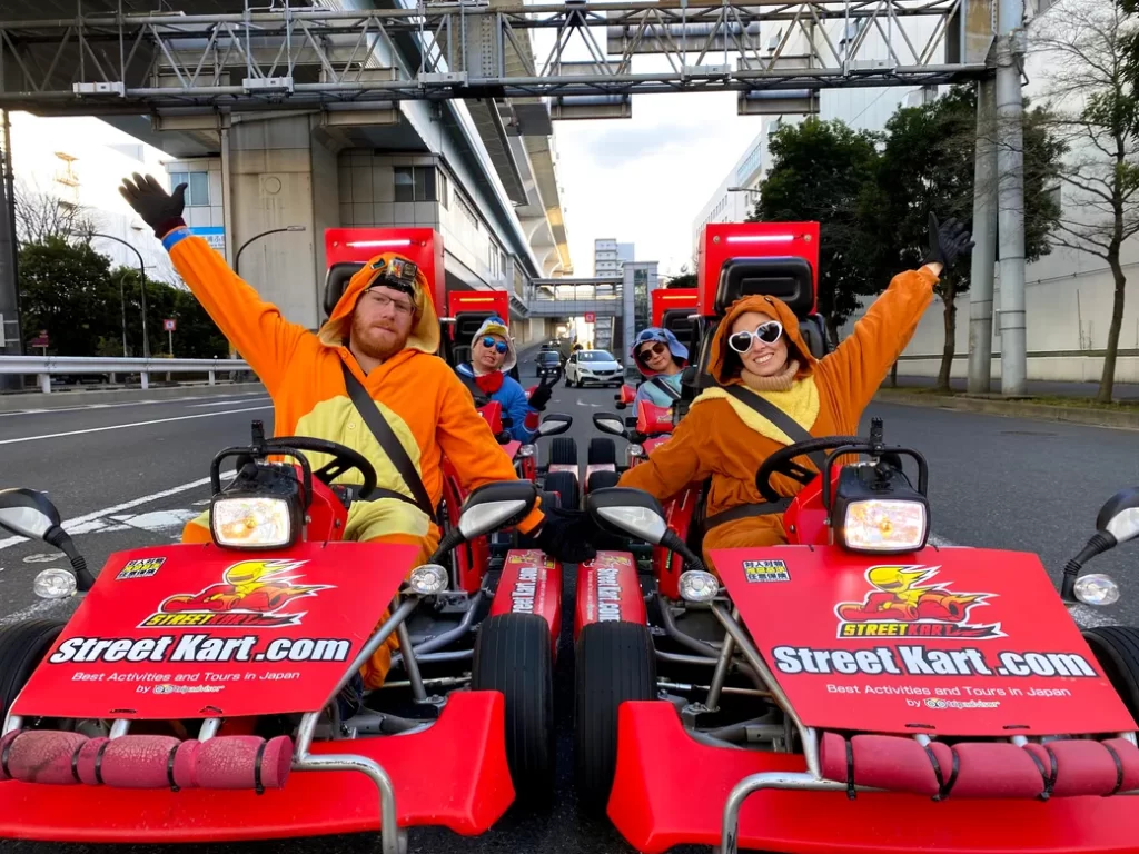 People in orange animal onesies driving red street go-karts on a city street, waving at the camera.