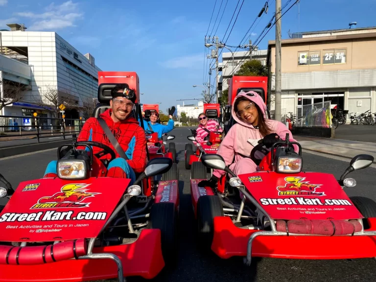 Friends in colorful costumes drive red Street Kart go-karts on a city street, smiling at the camera.