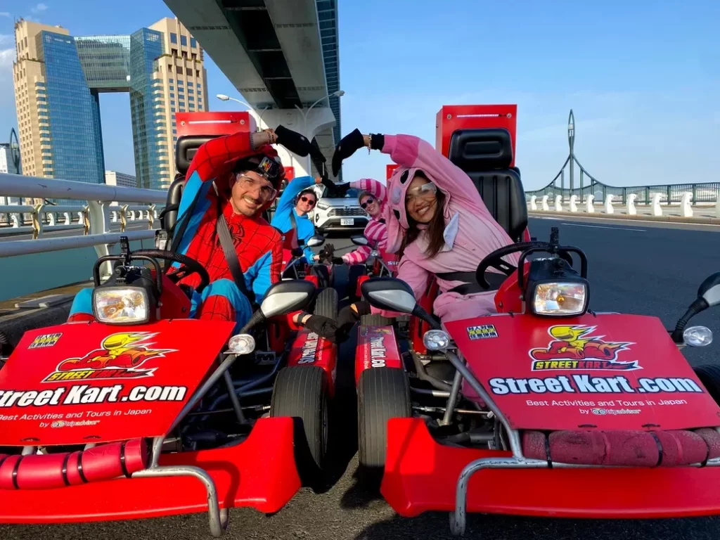 Group of friends in Spider-Man and pink bunny costumes posing in red go-karts on a bright city bridge, smiling at the camera on a street kart ride with Tokyo-like skyline in background.