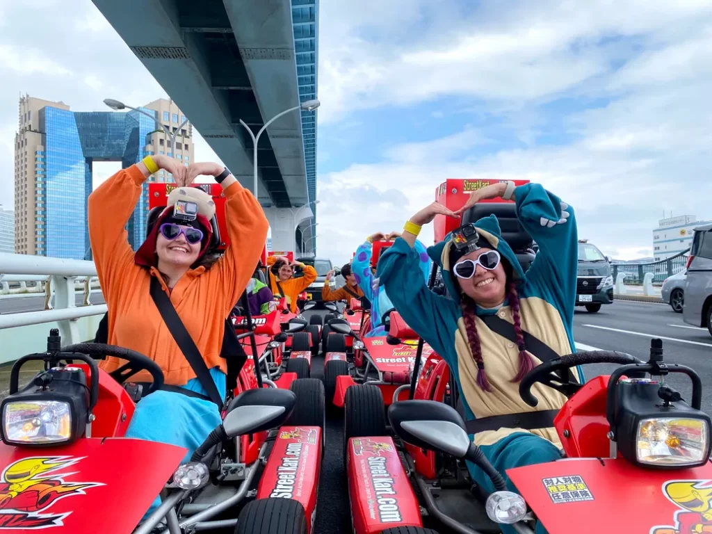 Group of people in red go-karts on a bridge, making heart shapes with their arms and smiling at the camera, wearing colorful outfits and GoPro head mounts.