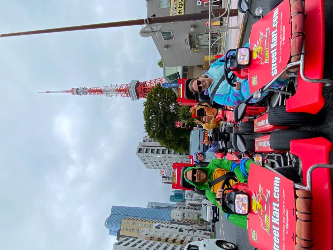 People ride bright red go-karts in a city parade, wearing colorful jackets with a tall tower and skyscrapers in the background.